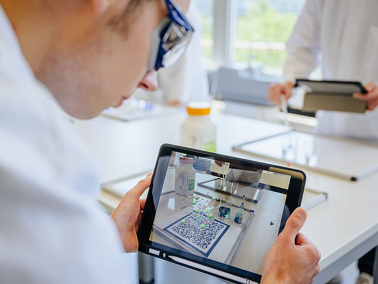 Ein Chemie Student betrachtet einen Experimentaufbau durch ein Ipad, welches dem Augmented Reality Elemente auf den Aufbau projiziert.