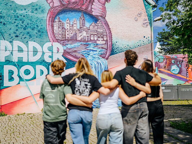 Fünf Studierende laufen Arm in Arm auf eine mit Graffiti gestaltete Wand zu, auf der der Schriftzug Paderborn zu lesen ist. 