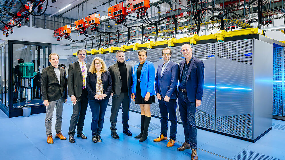 Fünf Männer und zwei Frauen, alle elegant gekleidet, stehen in einem Rechenzentrum und lächeln.