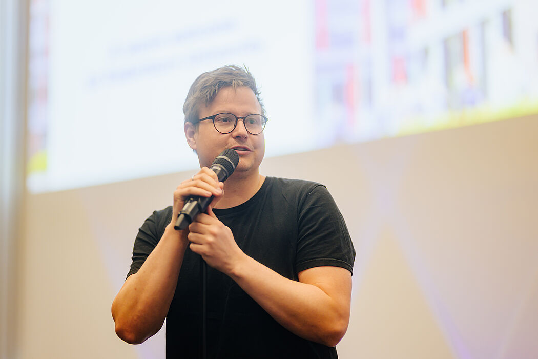 Ein Mann im schwarzen T-Shirt hält ein Mikrofon in der Hand und spricht.