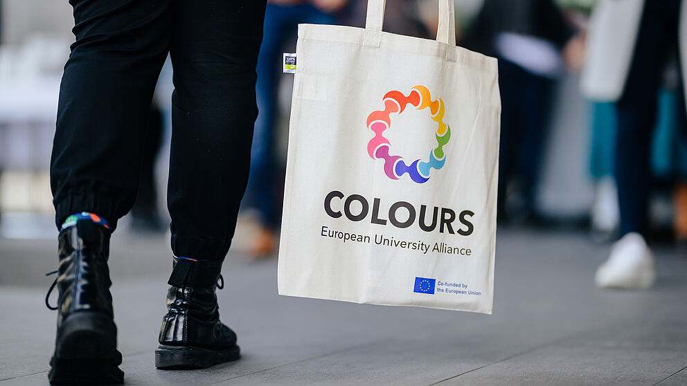 Beine in schwarzer Hose und mit schwarzen Stiefeln, daneben eine Tasche mit Aufdruck des Logos der europäischen Hochschulallianz COLOURS. 